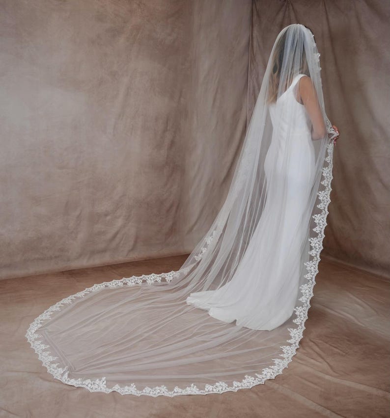 K&ouml;nnte beinhalten: Ein langer, wei&szlig;er Brautschleier mit zarter Spitzenborte. Der Schleier ist &uuml;ber einem wei&szlig;en Kleid drapiert und erzeugt einen flie&szlig;enden, eleganten Look. Die Spitzenborte verleiht dem Brautaccessoire einen Hauch von Raffinesse.