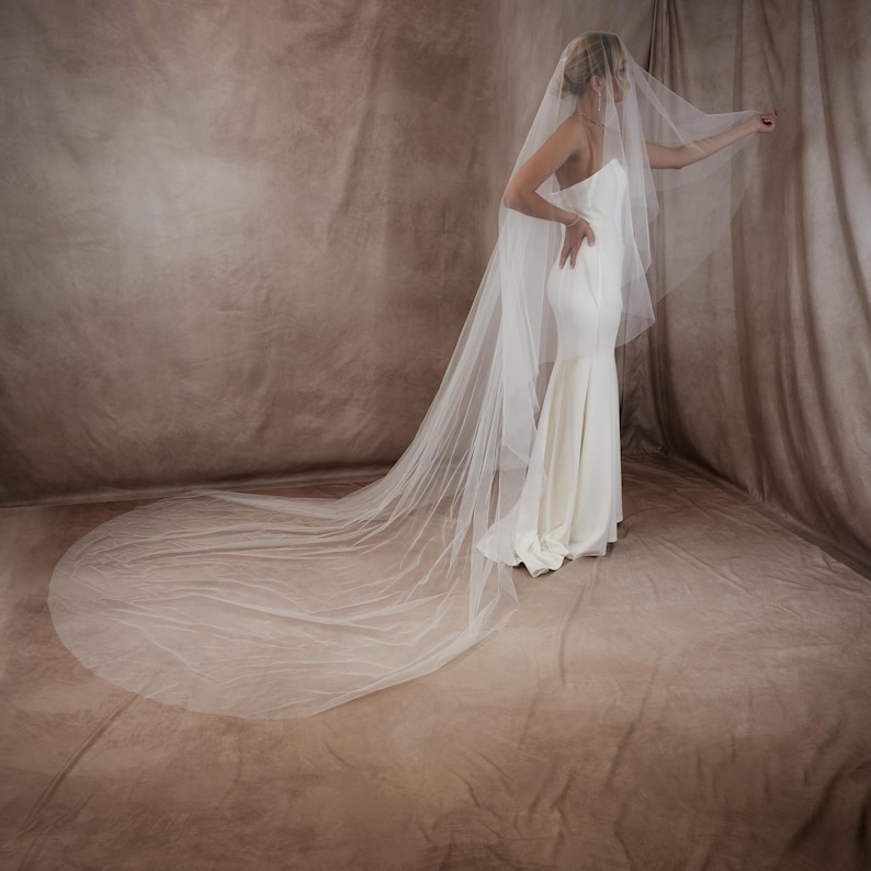 May include: A floor-length white wedding veil made of sheer, lightweight tulle fabric. The veil features a long, flowing train that extends across the floor, paired with a strapless, form-fitting white gown that reaches the ground.
