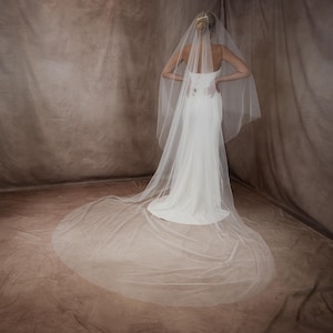 May include: A long, sheer white bridal veil made of fine tulle fabric, featuring a cascading design that extends into a floor-length train. The veil is attached to a hair accessory and drapes over a simple, floor-length white gown with thin straps.