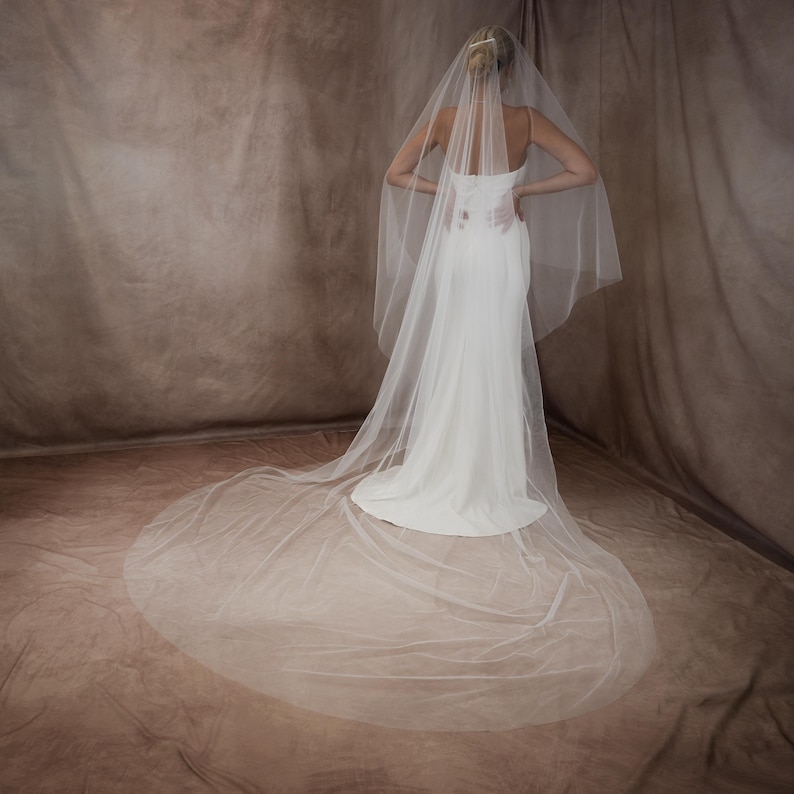 May include: A long, sheer white bridal veil made of fine tulle fabric, featuring a cascading design that extends into a floor-length train. The veil is attached to a hair accessory and drapes over a simple, floor-length white gown with thin straps.