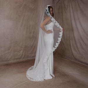 May include: A long, white wedding veil with a detailed lace edge. The veil is worn by a person wearing a white dress. The lace trim adds a touch of elegance to the bridal accessory.