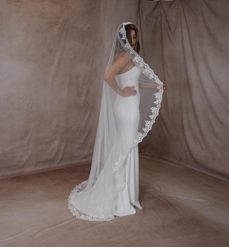 May include: A long, white wedding veil with a detailed lace edge. The veil is worn by a person wearing a white dress. The lace trim adds a touch of elegance to the bridal accessory.