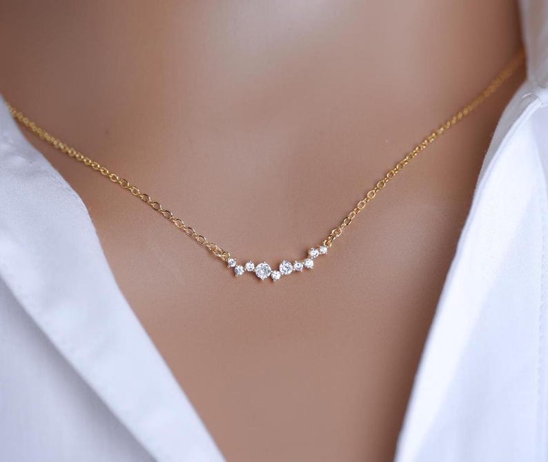 May include: A delicate gold chain necklace featuring a horizontal cluster of small, clear, round-cut stones arranged in a graduated, asymmetrical pattern. The stones are set in a bright, polished metal finish, creating a subtle, sparkling focal point.