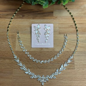 May include: A gold-toned bridal jewelry set featuring a necklace, bracelet, and earrings. The necklace and bracelet have a leaf-like design with clear crystals. The earrings are dangling, with a similar crystal design. The set is displayed on a wooden surface.