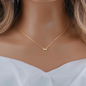May include: A delicate gold chain necklace featuring a single, small, rectangular baguette-cut clear stone pendant centered on the chain. The necklace has a thin, polished gold-tone link design that rests against the collarbone area.