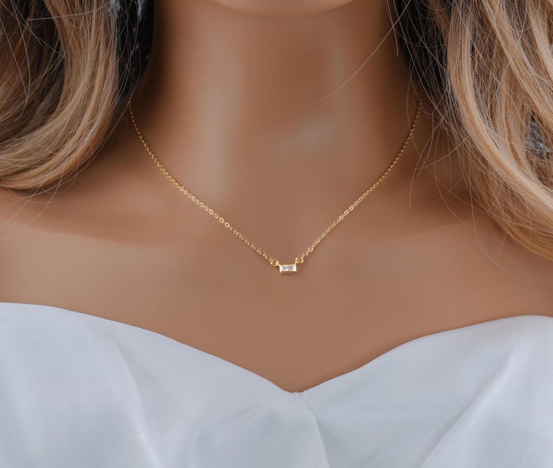 May include: A delicate gold chain necklace featuring a single, small, rectangular baguette-cut clear stone pendant centered on the chain. The necklace has a thin, polished gold-tone link design that rests against the collarbone area.