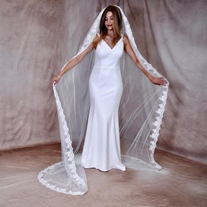 K&ouml;nnte beinhalten: Ein wei&szlig;es Brautkleid mit V-Ausschnitt und langem, flie&szlig;endem Rock. Die Braut tr&auml;gt einen langen, durchsichtigen Schleier mit zarter Spitzenborte. Der Schleier reicht bis zum Boden und sorgt f&uuml;r einen eleganten und romantischen Look.