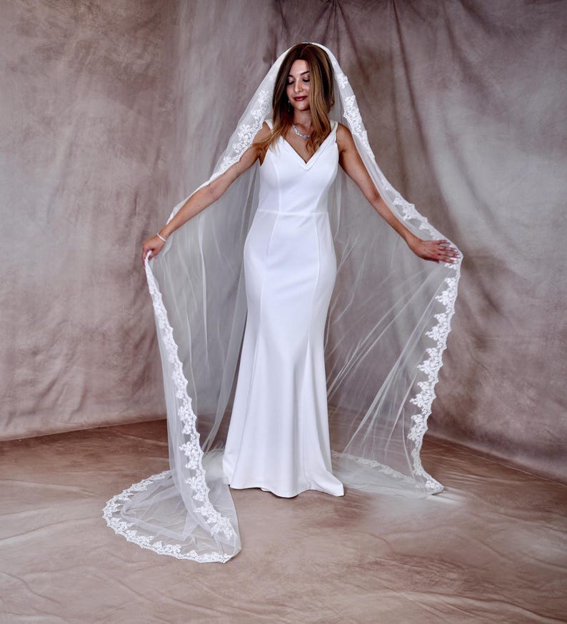 K&ouml;nnte beinhalten: Ein wei&szlig;es Brautkleid mit V-Ausschnitt und langem, flie&szlig;endem Rock. Die Braut tr&auml;gt einen langen, durchsichtigen Schleier mit zarter Spitzenborte. Der Schleier reicht bis zum Boden und sorgt f&uuml;r einen eleganten und romantischen Look.