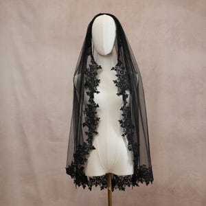 Puede incluir: Un velo negro de tul transparente con intrincados bordados de encaje floral negro a lo largo de los bordes. El velo est&aacute; colocado sobre un maniqu&iacute; blanco, mostrando el detallado ribete de encaje festoneado que bordea toda la tela.