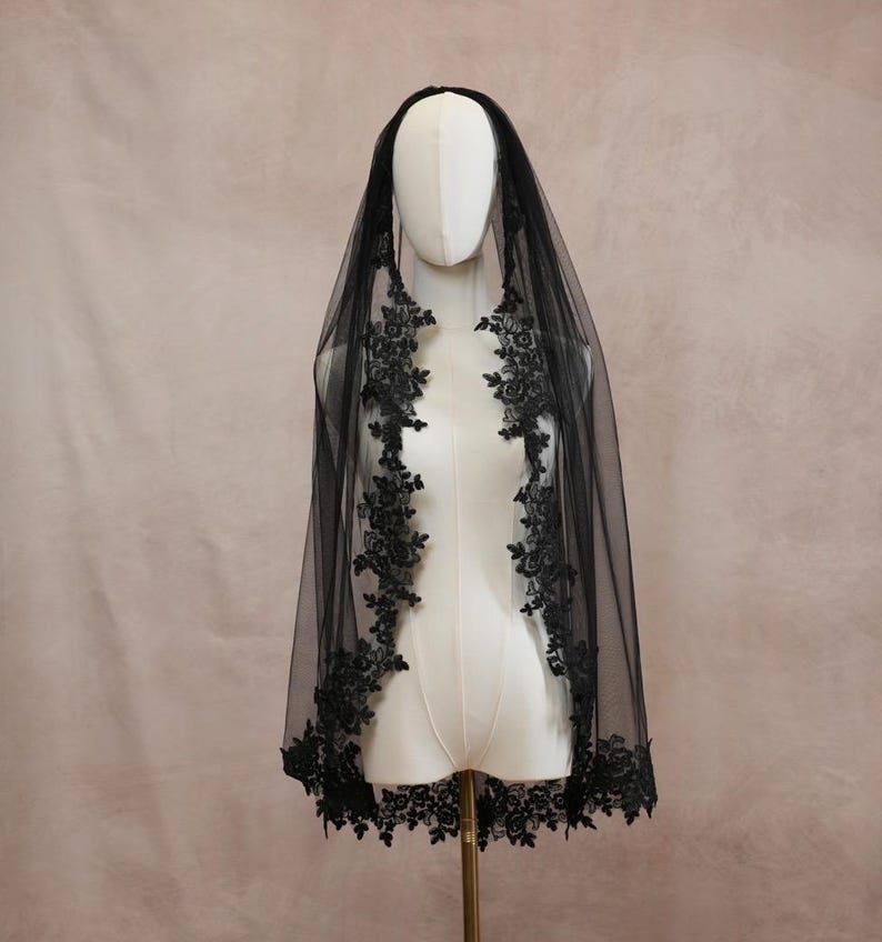 Puede incluir: Un velo negro de tul transparente con intrincados bordados de encaje floral negro a lo largo de los bordes. El velo est&aacute; colocado sobre un maniqu&iacute; blanco, mostrando el detallado ribete de encaje festoneado que bordea toda la tela.