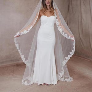 May include: A white bridal gown with a mermaid cut and a strapless bodice. The bride is wearing a long, sheer veil with delicate lace trim. The veil is draped over the shoulders, creating an elegant look.