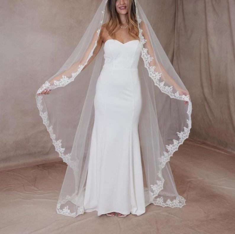 May include: A white bridal gown with a mermaid cut and a strapless bodice. The bride is wearing a long, sheer veil with delicate lace trim. The veil is draped over the shoulders, creating an elegant look.