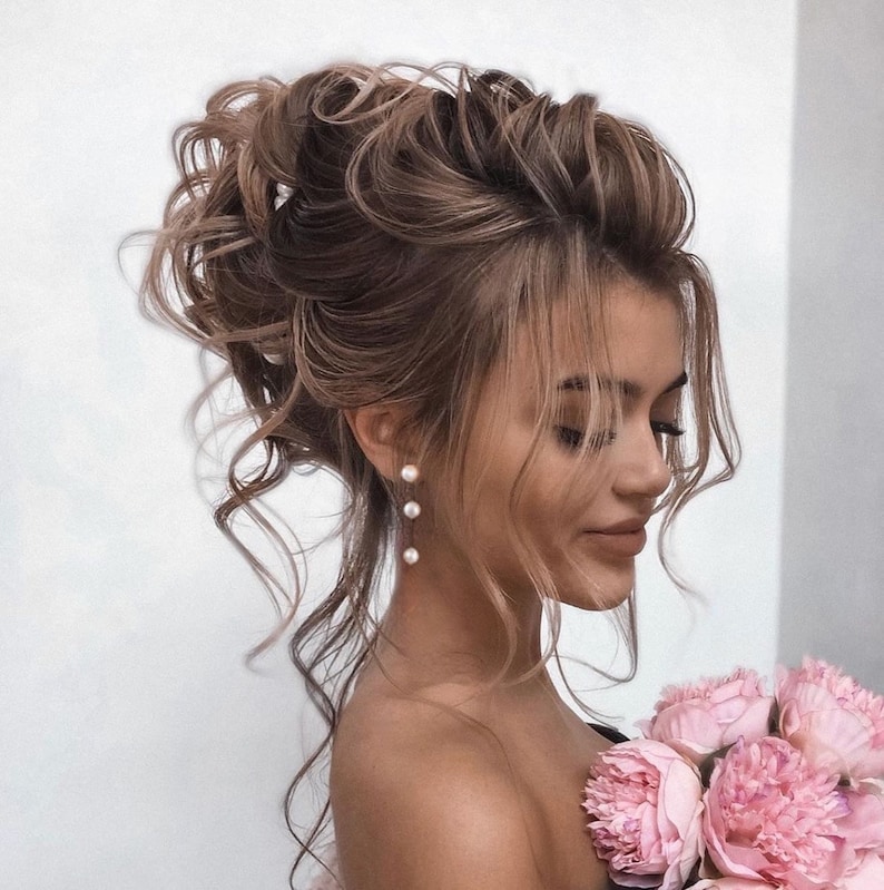 May include: A woman with long, brown hair styled in a high bun with loose curls. She is wearing pearl earrings and has a bouquet of pink peonies in her hand.