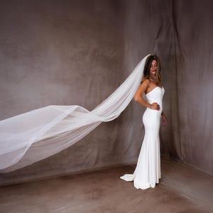 May include: A floor-length white strapless gown with a fitted bodice and a long, flowing sheer white veil that extends horizontally across the frame. The dress features a clean, minimalist silhouette that flares slightly toward the floor hemline.