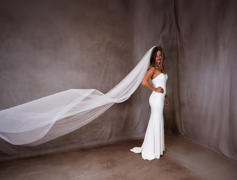 May include: A floor-length white strapless gown with a fitted bodice and a long, flowing sheer white veil that extends horizontally across the frame. The dress features a clean, minimalist silhouette that flares slightly toward the floor hemline.