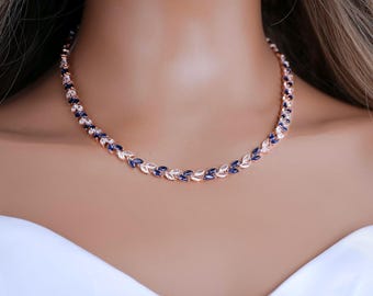 May include: A rose gold tone necklace featuring a repeating leaf-like pattern set with alternating marquise-cut dark blue and clear white stones. The metal chain links the stones in a continuous vine design that rests around the neck area.