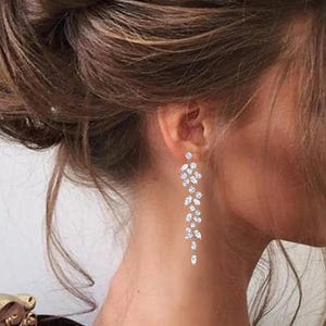 May include: A long, silver dangle earring with a cascade of clear, sparkling stones. The earring is worn by a person with long, brown hair.