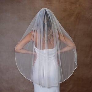 May include: A sheer white bridal veil made of fine tulle fabric, featuring a delicate scalloped edge adorned with small, evenly spaced white beads. The veil is draped over the head and shoulders, falling in soft, translucent layers down the back.