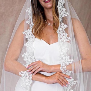 May include: A white bridal veil featuring delicate floral lace edging. The veil is worn over a white dress, creating a soft, romantic appearance. The lace detailing adds a touch of elegance to the bridal ensemble.