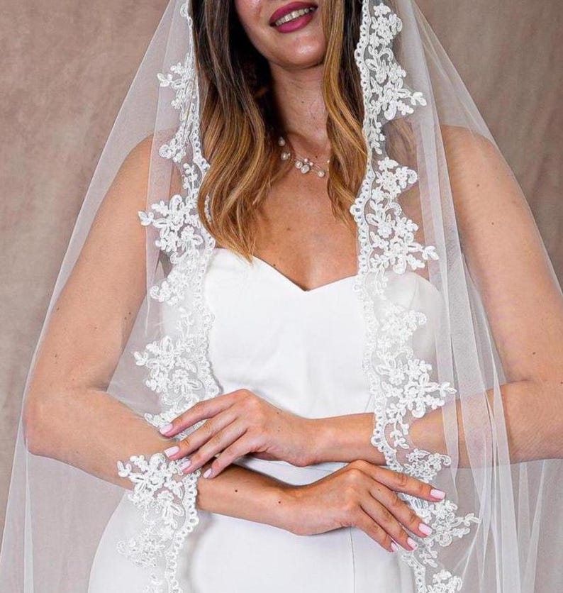 May include: A white bridal veil featuring delicate floral lace edging. The veil is worn over a white dress, creating a soft, romantic appearance. The lace detailing adds a touch of elegance to the bridal ensemble.