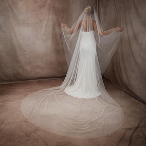 May include: A long, sheer white bridal veil made of fine tulle fabric, featuring a floor-sweeping train that extends behind the wearer. The veil is attached to the hair and drapes over the shoulders, creating a soft, translucent layered effect.