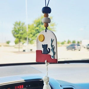 May include: A white ceramic car air freshener with a black cat silhouette, a yellow sun, and green leaves. The air freshener is hanging from a black string with brown, white, and black beads.