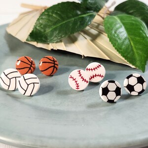 May include: Six pairs of small, round earrings featuring different sports ball designs. The earrings are white with red, black, and orange details. The designs include a basketball, baseball, football, and volleyball.