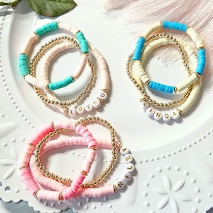 May include: Three sets of beaded bracelets in pink, cream, and turquoise. Each set includes a gold beaded bracelet and a bracelet with flat, colourful beads. One bracelet spells "LOVE U", another "LINDA", and the third "RILEY".