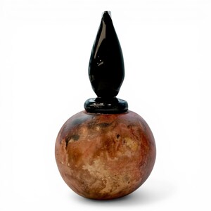 May include: A decorative bottle with a round, reddish-brown base and a black, teardrop-shaped stopper. The bottle has a textured surface and a small black ring at the base of the stopper.
