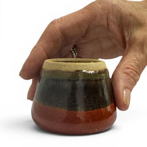 May include: A small, tapered ceramic cup featuring a wide, unglazed beige rim and a smooth, glossy body. The exterior displays horizontal bands of colour, transitioning from a dark olive green at the top to a deep black middle and a reddish-brown base.