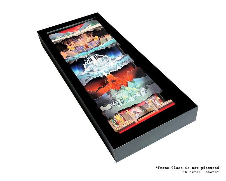 May include: A framed layered paper art piece depicting a scene with a waterfall, a mountain, a forest, and a building. The artwork is in a black frame and is displayed on a white background.