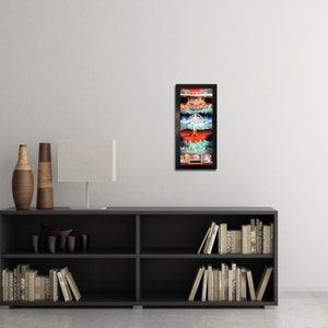 May include: A framed print of a colorful, layered illustration of a city skyline. The print is in a black frame and is hanging on a white wall above a black bookshelf.