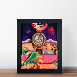 Könnte beinhalten: Ein mehrschichtiges Schattenbox-Kunstwerk in einem schwarzen Rahmen zeigt eine Figur mit grüner Kapuze auf einer Holzplattform. Die Szene stellt einen Stadtplatz mit einem großen Uhrturm, einem roten Mond mit Gesicht am Himmel und Feuerwerk dar.