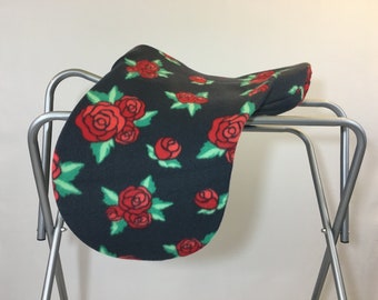 May include: A black saddle pad featuring a floral design of red roses and green leaves. The pad is made from a soft, fleece material and is shown on a silver metal stand.