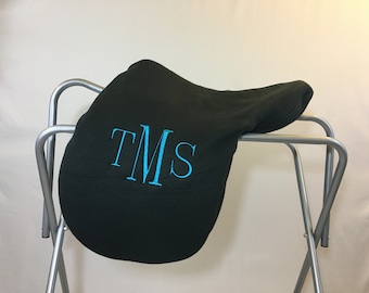 May include: A black fleece saddle pad with the monogram "TMS" embroidered in turquoise blue.