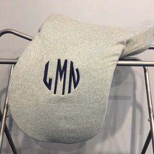 Puede incluir: Un cojín de silla de montar de vellón gris con un monograma azul marino de las letras LMN en un círculo.