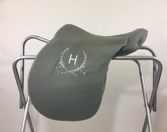 May include: A grey fleece saddle pad with a white embroidered wreath and the letter "H" in the centre.