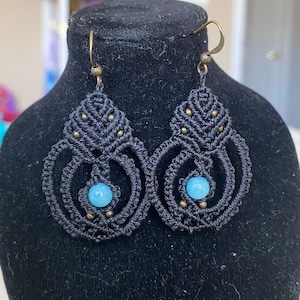May include: A pair of black macrame earrings with blue gemstone accents. The earrings are made with a delicate knotting technique and feature a teardrop shape.