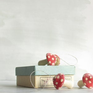 May include: A set of three red and white polka dot mushroom ornaments with a white string attached to each. The ornaments are sitting on top of a light blue box with a white label that says "No. 4412 Ltd. TONS".