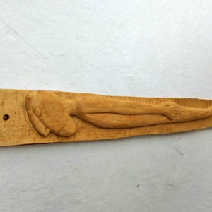 Può includere: Una scultura a rilievo in legno intagliato raffigurante una figura umana reclinata. La figura è intagliata in legno marrone chiaro e si trova su una base rettangolare di legno. La scultura è montata su un muro bianco.