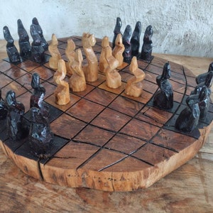 Puede incluir: Juego de ajedrez de madera tallada a mano. El tablero es una losa de madera natural circular con una cuadrícula de cuadrados. Las piezas de ajedrez están estilizadas, con tonos negros y madera natural. Un juego único, artesanal.