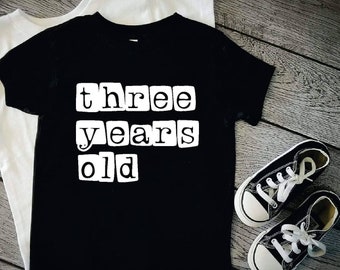 May include: Black t-shirt with white text that reads "three years old" in a blocky font. The shirt is laid out on a wood surface with a white t-shirt and black sneakers beside it.