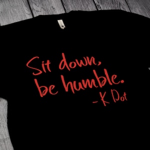 May include: Black t-shirt with the red text "Sit down, be humble. - K Dot." The shirt has a crew neck and a tag at the neckline. The text is in a handwritten style.