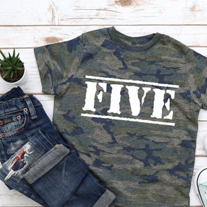 May include: A camouflage print t-shirt with the word "FIVE" printed in white on the front. The shirt is laid out on a white wooden surface with a pair of blue jeans and two pairs of light blue sneakers.