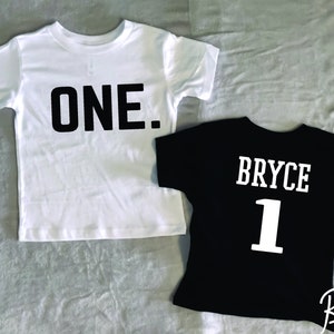 May include: A white short-sleeved t-shirt with the word ONE. printed in black block letters and a black short-sleeved t-shirt with the name BRYCE and the number 1 printed in white block letters on the front of the garments.