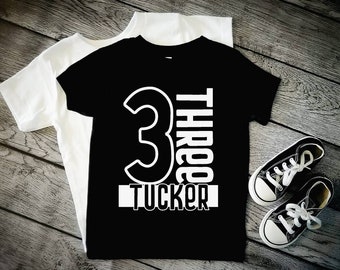 May include: Black t-shirt with white text that says "Three Tucker" and a large white number "3" on the front.