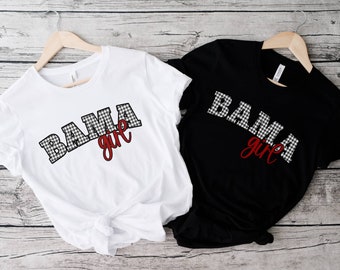 May include: A white t-shirt and a black t-shirt, each featuring the text BAMA in a black and white houndstooth pattern with the word girl written in red cursive script below the letters. Both shirts are displayed on wooden hangers.