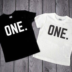May include: A black short-sleeved t-shirt with the word ONE. printed in white and a white short-sleeved t-shirt with the word ONE. printed in black. Both shirts feature a simple crew neckline and a standard fit for casual wear.