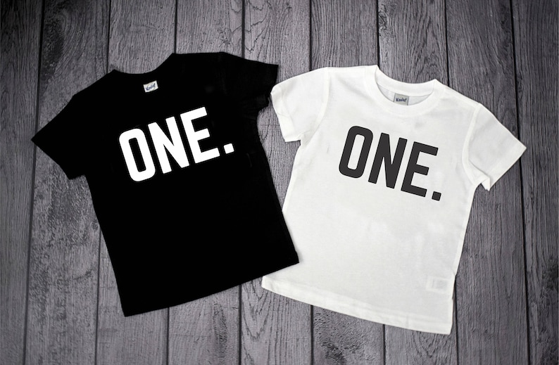 May include: A black short-sleeved t-shirt with the word ONE. printed in white and a white short-sleeved t-shirt with the word ONE. printed in black. Both shirts feature a simple crew neckline and a standard fit for casual wear.
