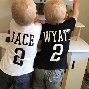 May include: Two short-sleeved t-shirts, one white with black text reading JACE and the number 2, and one black with white text reading WYATT and the number 2. Both shirts feature bold, block-style lettering and numerals printed on the back.